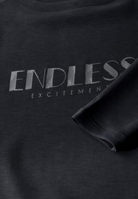 Mustapaita, jossa on metallinhohtoisella hopealla kirjoitettu "ENDLESS EXCITEMENT". Sileä kangas, pyöreä pääntie ja hihansuut on käännetty.