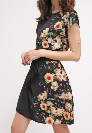 Woman wearing a black short-sleeve dress with a floral pattern and a tied waist belt, standing against a plain light background.