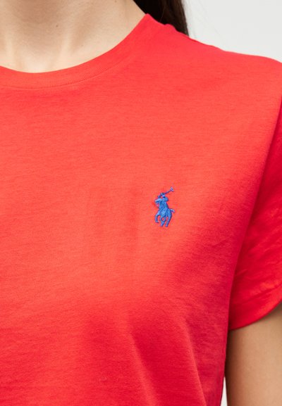 Κοντινό πλάνο ενός ατόμου που φοράει κόκκινο μπλουζάκι με μικρό μπλε κεντημένο λογότυπο παίκτη πόλο στο αριστερό στήθος.