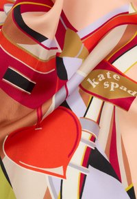 Hedvábný šátek s abstraktními geometrickými tvary v červené, béžové, černé a zlaté barvě, s nápisem "kate spade" na zlatém banneru.