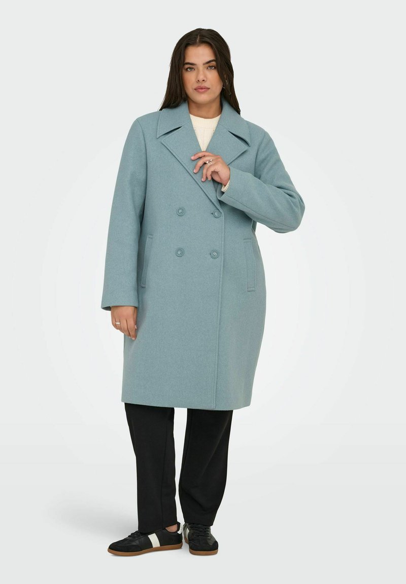 Gai zils vilnas maisījuma mētelis ar dubultu pogu slēgšanu, lieliem atlokiem un divām pogām, kombinēts ar melnajiem biksēm un melnajām sporta apavām.