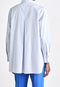 Světle modrá a bílá pruhovaná košile s knoflíky, se záhybem na zádech a dlouhými rukávy se zdrhovadly, vyrobená z hladké látky. Nositelná s modrými kalhotami.