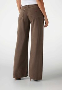 Pantalones de mezclilla marrón de pierna ancha con cintura alta, que cuentan con dos bolsillos traseros y una etiqueta de logo cerca de la cinturilla. Textura suave y acabado limpio.
