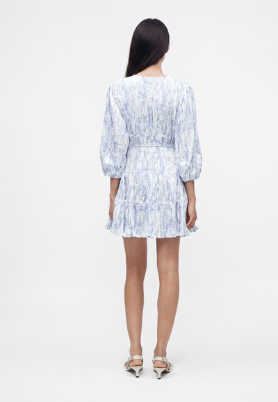 Femme aux longs cheveux bruns porte une robe courte bleu clair à motifs avec des manches bouffantes et des sandales blanches à talons, vue de dos.