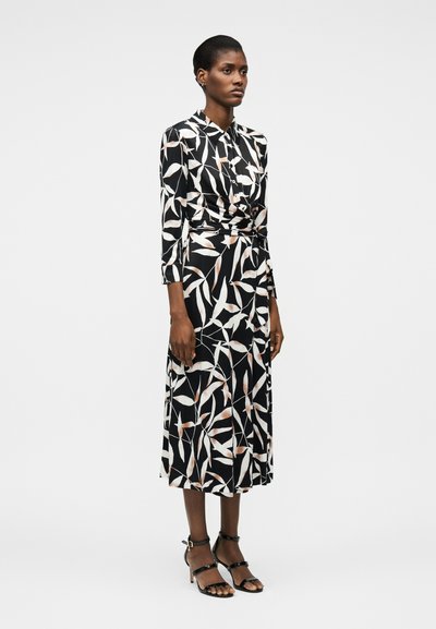 Γυναίκα στέκεται φορώντας μαύρο midi φόρεμα με λευκά και απαλό ροζ φύλλα, μακριά μανίκια και μαύρα σανδάλια με τακούνια.