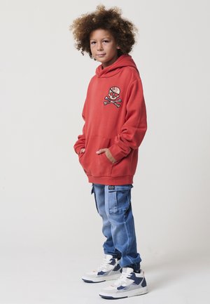 Bambino con capelli ricci che indossa un hoodie rosso con una toppa a forma di teschio, pantaloni cargo blu e sneakers bianche, in piedi con le mani nelle tasche.