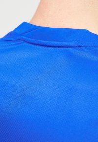 Κοντινό πλάνο του λαιμού και του άνω μέρους της πλάτης ενός ατόμου που φοράει πουκάμισο από φωτεινό μπλε υλικό με υφή και ραμμένη λεπτομέρεια γιακά.