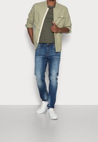 Camiseta verde oliva debajo de una chaqueta abotonada verde claro, combinada con jeans azules y zapatillas blancas. La chaqueta tiene un bolsillo en el pecho y broches.