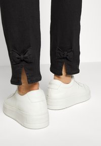 Calças pretas com detalhes de laço na bainha e fendas, combinadas com ténis brancos em pele com sola grossa.