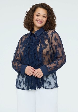 Uśmiechnięta kobieta o kręconych włosach, ubrana w granatową przezroczystą koronkową bluzkę z falbankami i białe spodnie, stojąca na jednolitym tle.