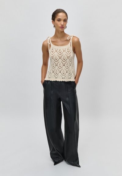 Γυναίκα που φοράει μπεζ πλεκτό αμάνικο τοπ με δεμένους ιμάντες στους ώμους και μαύρο δερμάτινο παντελόνι με φαρδιά πόδια, στέκεται με τα χέρια στις τσέπες.