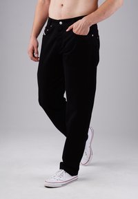 Schwarze Denim-Hose mit geradem Schnitt, ausgestattet mit Fronttaschen, metallischen Details und einem schlichten Design. Getragen mit weißen Sneakers.