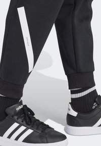 Underben iförda svarta träningsbyxor med vitt streck och svarta Adidas sneakers med vita ränder, i kombination med svarta och vita randiga strumpor.