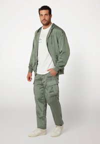 Világos zöld bomber dzseki kapucnival, amelyhez illeszkedő cargo nadrág tartozik zsebekkel. A modellen fehér póló és fehér sportcipő van.