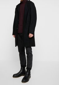 Musta villatakki nappisuljennuksella, tumma punainen ja tummaraidallinen poolopaita sen päällä, yhdistettynä mustiin farkkuihin ja mustiin nilkkureihin.