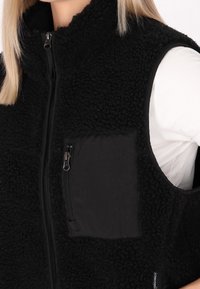 Veste sans manches en polaire noire avec fermeture éclair, col montant et poche poitrine zippée. Tissu texturé avec accents lisses.