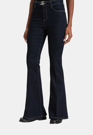 Donna che indossa jeans a zampa blu scuro con cuciture a contrasto e scarpe nere, in piedi davanti a uno sfondo bianco.