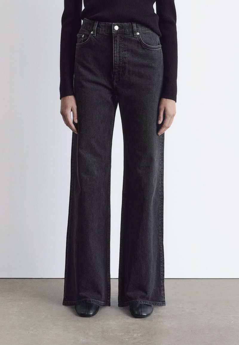 Jean en denim noir taille haute, à jambes larges, avec une finition légèrement texturée, comportant cinq poches et des boutons en métal.