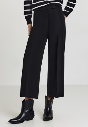 Pantalons larges noirs en tissu lisse, dotés d'une taille haute et de plis subtils, associés à des bottines noires.