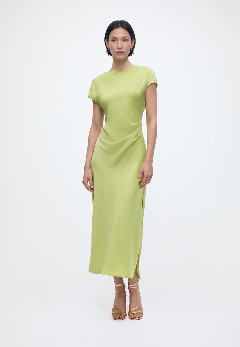 Femme debout, face à l'avant, portant une robe midi à manches courtes vert clair avec une fente latérale et des sandales à talons hauts beiges à lanières.