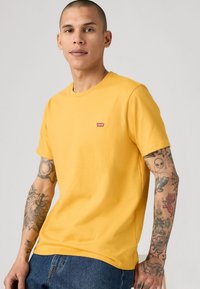 Jongeman met een geschoren hoofd die een geel Levi's T-shirt en blauwe jeans draagt, met gedetailleerde tatoeages op beide armen tegen een effen achtergrond.
