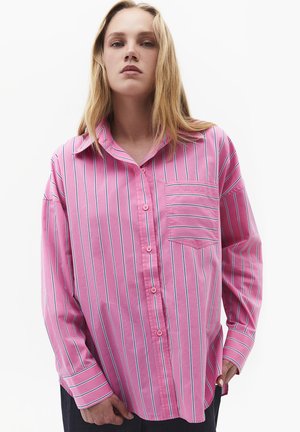 Junge Person mit langen blonden Haaren, die ein lockeres rosa Hemd mit vertikalen weißen und blauen Streifen und einer gestreiften Brusttasche trägt.
