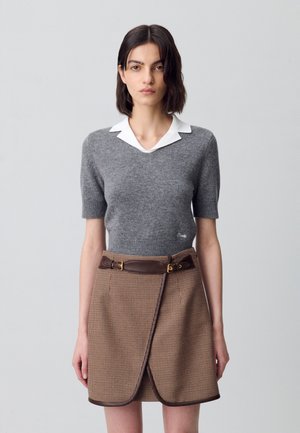 Una modella si trova davanti a uno sfondo grigio chiaro, indossando un maglione grigio a maniche corte e una gonna marrone a quadretti con una cintura larga.