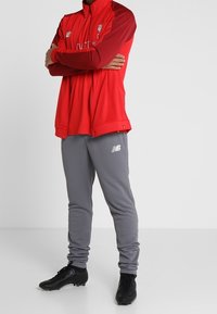 Rød lynlåsjakke med maroon detaljer, parret med grå sweatpants; sorte fodboldstøvler. Logo detaljer på jakke og bukser.