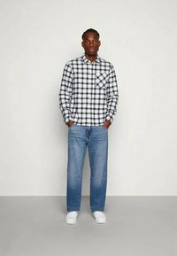 Ruudukujuline flanellsärk tumesinises ja valges, ees tasku, koos helesiniste teksade ja valgete kingadega, seismas neutraalses taustas.