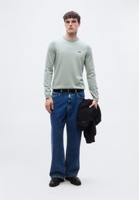 Νέος άνδρας όρθιος, φοράει ανοιχτοπράσινο πουλόβερ, μπλε τζιν, μαύρη ζώνη, μαύρα παπούτσια, κρατώντας μαύρο σακάκι στο δεξί χέρι μπροστά σε απλό λευκό φόντο.