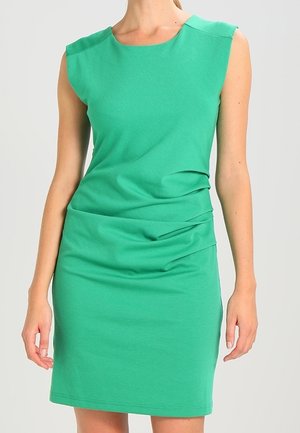 Ärmelloses, tailliertes grünes Kleid mit leichter Raffung auf der rechten Seite, getragen von einer stehenden Person mit entspannt an den Seiten hängenden Armen.