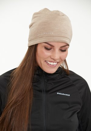 Beanie texturé beige avec un ajustement serré, présentant un motif rayé subtil et un logo blanc à l'avant. Une veste noire zippée est portée en dessous.