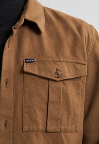 Bruin flanellen shirt met een knoopzak op de borst, spitse kraag en een geweven label. De stof lijkt zacht en duurzaam.
