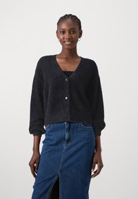 Črna puhasta jopica z V-izrezom in zapiranjem na gumb, z dolgimi rokavi in nabranimi manšetami, v kombinaciji z jeans krilom.
