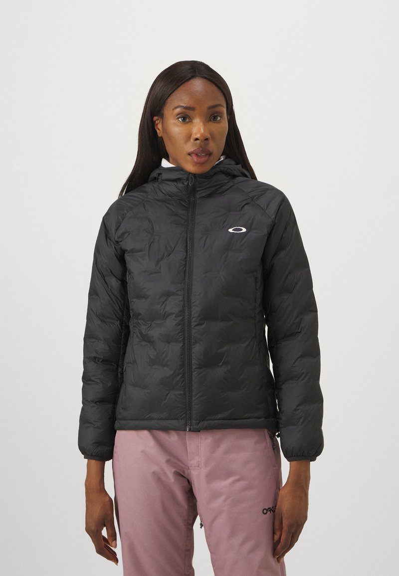 Schwarze Steppjacke mit Kapuze, Reißverschluss, gestepptem Design und Logo auf der Brust. Getragen mit hellrosa Hosen, die eine lockere Passform zeigen.