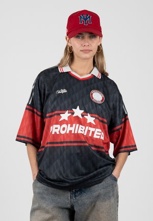 Joven que lleva una camiseta negra y roja de gran tamaño con el texto "PROHIBITED", una gorra roja con el logo azul "NY" y pendientes de aro, manos en los bolsillos.