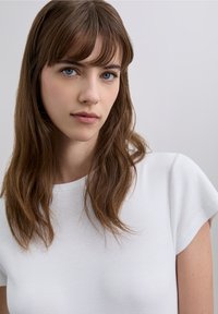 White ribbed t-shirt with a round neckline and short sleeves, worn by a person with long, wavy brown hair and natural makeup.