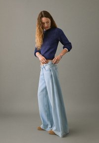 Svetlo modri široki kavbojke z visokim pasom, v kombinaciji s temno modrim pletenim puloverjem. Model prilagaja kavbojke, kar prikazuje njihov kroj.