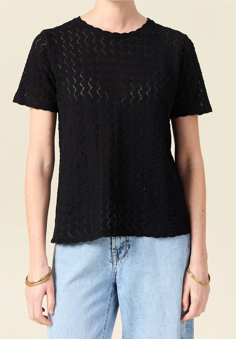 Haut en dentelle noir à manches courtes avec des bords festonnés, porté avec un jean bleu clair et des bracelets dorés aux deux poignets.