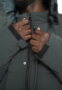 Grøn polstret jakke med høj krave, lynlås lukning og Velcro manchetter. Har en glat tekstur og metalliske trykknapper.