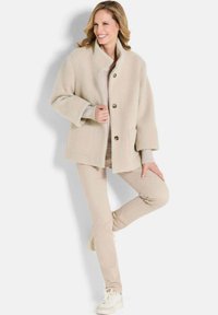 Beige textuur blazer met een staande kraag, frontknopen en twee zakken, gedragen over een lichte trui en beige broek, gecombineerd met witte sneakers.