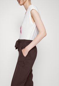 Top sem mangas branco com um gráfico rosa, combinado com calças elásticas de cintura alta castanhas escuras com um laço na cintura e bolsos laterais.