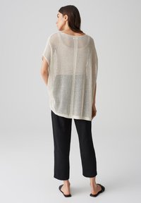 Pull crème en tricot ouvert avec une coupe ample, présentant un décolleté large et des manches tombantes. Associé à un pantalon noir à jambes droites et des sandales.