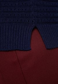 Granatowa dzianina z ażurowym wzorem i prążkowanym wykończeniem, zestawiona z gładkim bordowym materiałem. Posiada niewielki rozcięcie przy krawędzi.