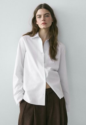 Frau mit langen braunen Haaren, die ein locker sitzendes weißes Hemd mit Knöpfen und dunkelbraune Hosen vor einem schlichten hellgrauen Hintergrund trägt.