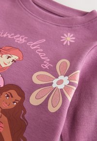 Lilla sweatshirt med tegneseriefigurer og et stort glitrende blomsterprint, med teksten "Princess dreams" i kursiv. Blød tekstur.