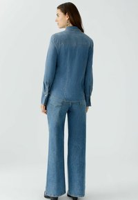 Denimhemd en wijde jeans in een middenblauwe wassing. Het shirt heeft een puntkraag, lange mouwen en stikdetails; de jeans hebben achterzakken.