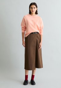 Junge Frau trägt ein apricotfarbenes Sweatshirt, einen braunen Midirock mit einem schwarzen Gürtel, rote Socken und schwarze Schuhe, steht vor einem schlichten hellgrauen Hintergrund.