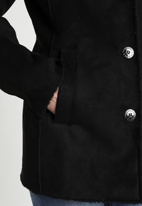 Czarna zamszowa kurtka o gładkiej strukturze, z srebrnymi guzikami i bocznymi kieszeniami. Krój ma lekko luźne dopasowanie.