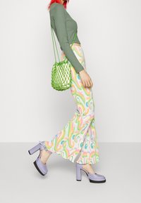 Grünes langärmliges Oberteil, bunt gemusterte Hosen mit Spiralen, lilafarbene Plateauschuhe und eine transparente grüne Bucket Bag mit geflochtenem Design.